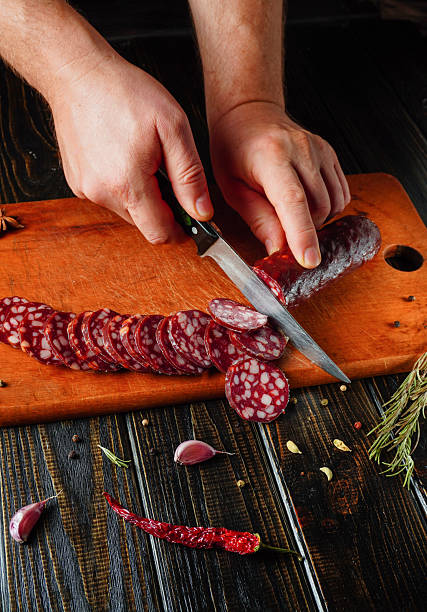 Hands carefully slice through salami on a wooden board, showcasing fresh ingredients and a warm, inviting kitchen atmosphere. The scene captures culinary art and preparation