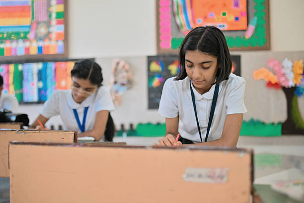 In a bright classroom decorated with vibrant student artwork, girls in white uniforms work intently at low desks, shading their pictures with colored pencils; the foreground student refines a bold, colorful composition while classmates in the background continue sketching, highlighting creativity, focus, and hands-on visual arts learning.