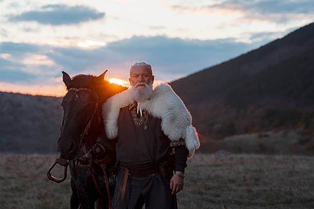 istock Elderly Viking man with a white beard, dressed in traditional attire, standing confidently beside a horse at sunset, set against a scenic landscape. 2246945889