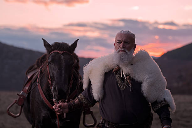 istock Elderly Viking man with a white beard, dressed in traditional attire, standing confidently beside a horse at sunset, set against a scenic landscape. 2246946020