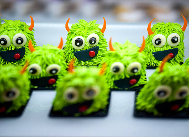 Close-up of whimsical green monster cakes topped with shaggy frosting, big eyes, orange horns, and cheeky smiles, displayed in neat rows for a fun bakery treat.