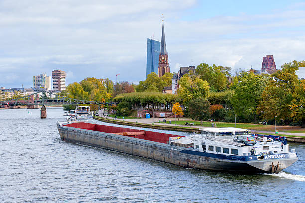 Frankfurt, Germany - October 21, 2025: Cargo vessel 'ANDIAMO' traverses the Main River, showcasing the city's blend of modern architecture and historic structures, with autumn trees lining the bank.