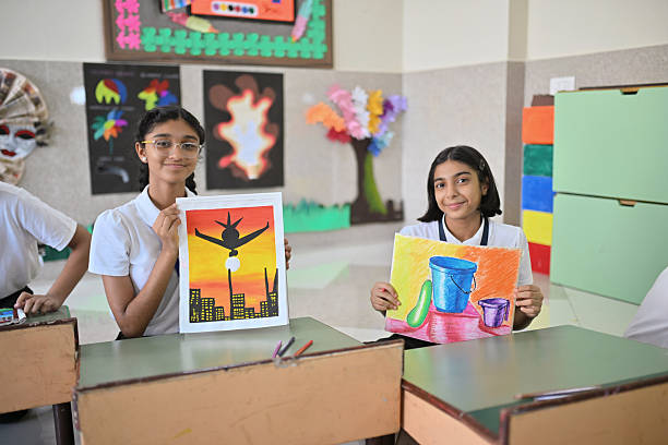 In an art room lined with student displays, two girls smile as they hold up their creations—one a bold sunset cityscape with an airplane silhouette, the other a bright still-life of a bucket, cup, and fruit. Their confident poses and vivid pieces showcase creativity, skill, and the joy of sharing their work.