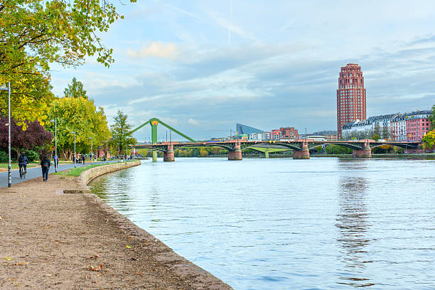 Frankfurt, Germany - October 21, 2025: Frankfurt's scenic Main River embankment features pedestrians and cyclists, providing views of historical bridges and the modern Westhafen Tower.
