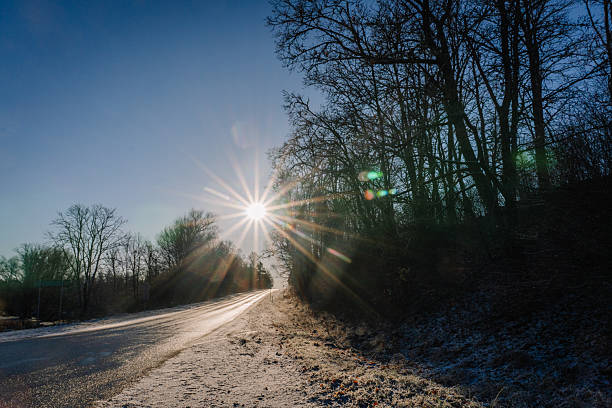 Sunlight beams over a snowy road, framed by trees, creating a tranquil winter atmosphere with a clear blue sky and bright reflections enhancing the beauty of nature