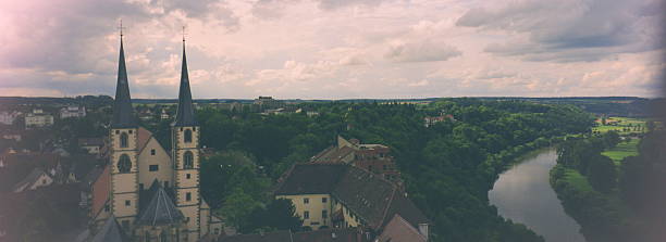 Stunning panoramic aerial view of Bad Wimpfen, a medieval spa town in Baden-Württemberg, Germany. The twin spires of the Evangelische Stadtkirche (Protestant Town Church) stand tall amid red-roofed half-timbered houses, with the serene Neckar River winding through dense green forests under a dramatic pink-tinged cloudy sky at sunset. Captures the timeless historic charm in a sweeping vista.