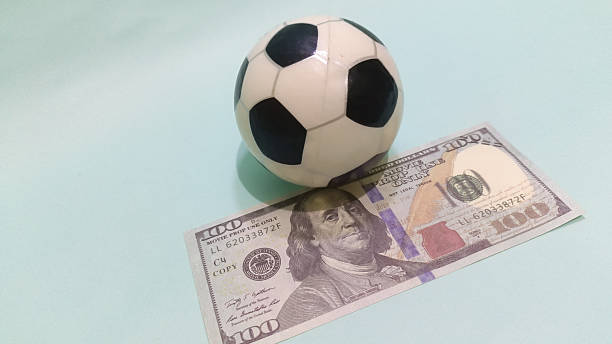 istock Soccer ball sits atop a one hundred dollar bill on a pale blue surface representing the intersection of sports. 2246963171