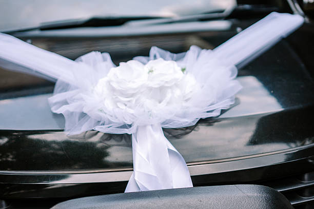 A dark wedding car is beautifully adorned with elegant white ribbons and a delicate floral arrangement, ready for a special celebration.