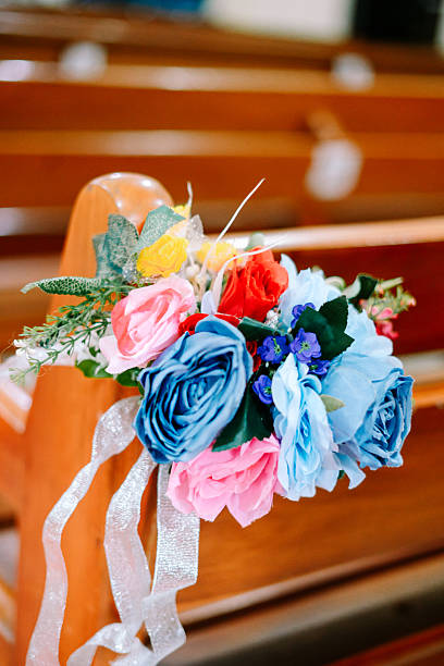 A vibrant artificial flower bouquet with pink, blue, red, and yellow roses beautifully adorns a wooden church pew, perfect for wedding or event decor.