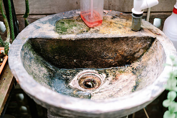 An old, weathered concrete sink basin, covered in moss and dirt, with a simple plastic faucet and a red soap dispenser. This rustic outdoor feature shows signs of age and natural decay.