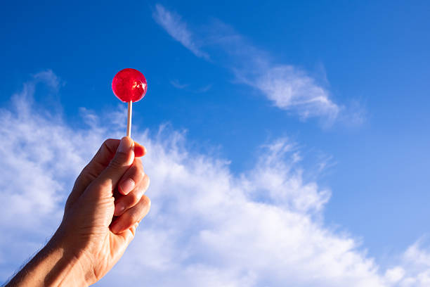 Hand holding a vibrant red lollipop against a backdrop of clear blue sky with soft white clouds, symbolizing childhood, sweetness, and simple pleasures in warm weather