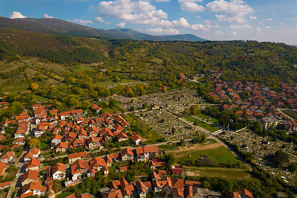 Aerial view displaying a rural balkan village with houses, green hills, and a large cemetery