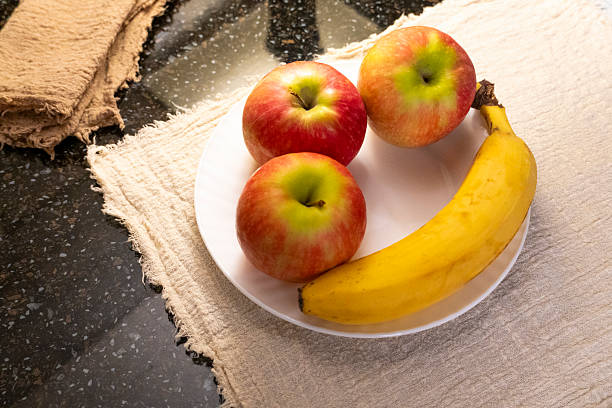 A white plate with red apples and a ripe banana on a beige placemat, accompanied by crispbread and a mug. Cozy kitchen setup ideal for healthy eating themes