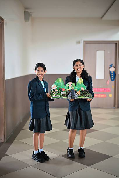 Two girls holds a detailed diorama featuring green mountains, stepped paths, and small figurines; indicating their school project, the bright craft contrasts with the neutral hallway, highlighting their teamwork and creativity.