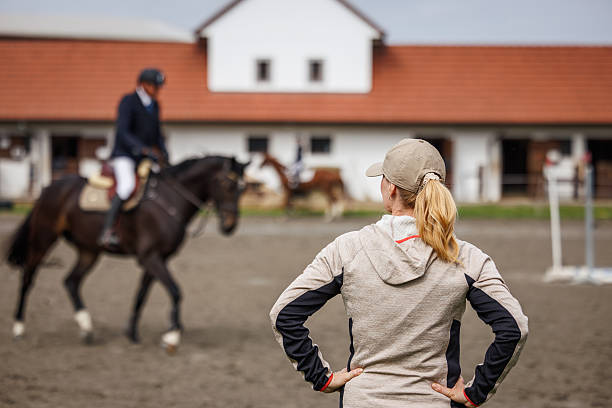 Female animal trainer supervising rider training before equestrian show jumping in paddock