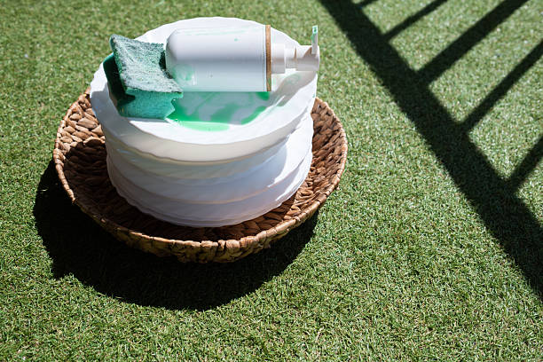 Stacked white ceramic dishes sitting in a wicker basket on green artificial grass, with a sponge and dish soap bottle on top, implying outdoor cleaning or a picnic cleanup