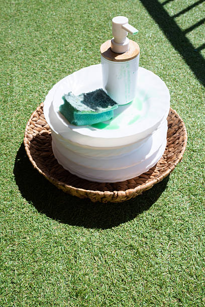 Stack of dirty white plates with green dish soap, scrub sponge and pump on a wicker tray set on vibrant artificial grass, evoking outdoor chores, summer cleanup and eco-friendly living