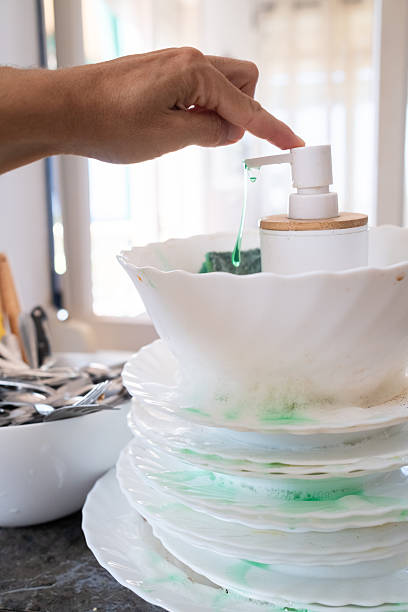 Hand pushing down a white soap dispenser, releasing green dishwashing liquid over a stack of dirty white plates and a bowl with a green sponge inside, highlighting essential house chores