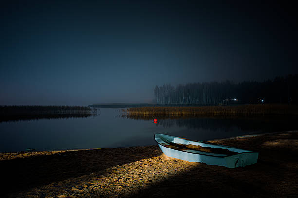 Atmospheric night photograph capturing a small, solitary boat resting on a sandy beach in Raisio, Southwest Finland. The scene is lit by an unseen light source, casting strong shadows and highlighting the texture of the boat and sand against the dark night. This image evokes feelings of solitude, mystery, and peaceful coastal tranquility, ideal for themes related to Nordic travel, nostalgia, and quiet evening scenes.