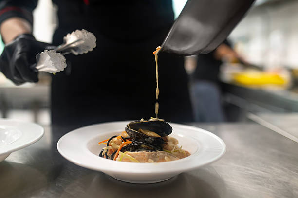Chef preparing pasta dish with mussels, vegetables, and sauce in professional kitchen