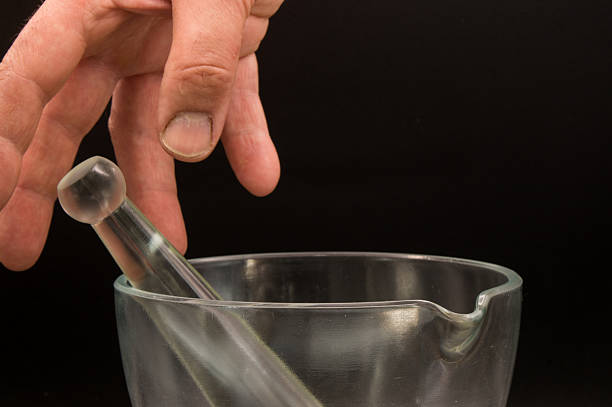 Action in a compounding pharmacy. A hand reaches down toward the glass pestle resting in a clear glass mortar, ready to begin grinding ingredients. Use for: pharmaceutical preparation, dosage mixing, herbalism, and scientific research.
