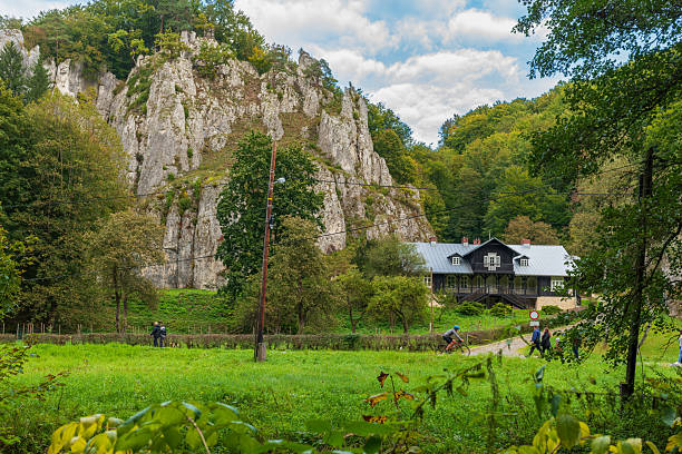 Ojcow National Park, Poland – October 5, 2025: A towering limestone cliff rises over a historic wooden house nestled among vibrant autumn trees. Visitors and a cyclist add life to the peaceful valley scene.