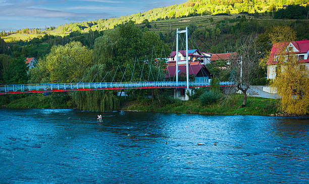 Light pedestrian suspension bridge stretching across a calm river, with fishermen standing in the shallow water below. Soft evening light, riverside houses, and lush greenery create a peaceful outdoor scene perfect for travel and lifestyle themes.