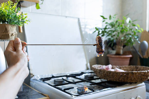 Person's hand holding a skewer with a marshmallow roasting over a kitchen gas stove flame, preparing a sweet treat with a burnt texture and creating a warm, cozy atmosphere at home