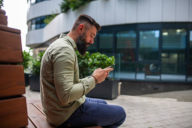 istock Bearded young man typing on phone outdoors 2247025673
