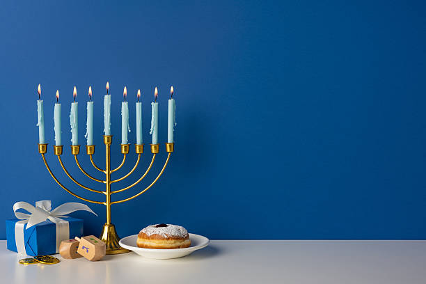 istock Blue wall backdrop frames a golden menorah with lit candles, a wrapped gift, dreidel, coins, and a donut on a plate, capturing warm festive light and Hanukkah spirit 2247028491