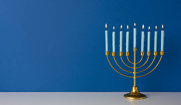 A golden menorah with nine lit candles sits on a white surface before a deep blue wall, evoking Hanukkah tradition, warmth, and holiday celebration