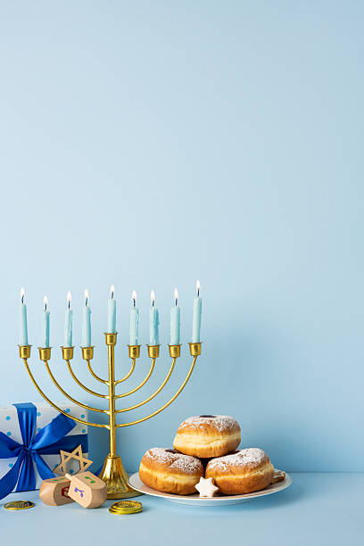 istock A vertical festive Hanukkah scene featuring a glowing menorah, powdered donuts, a plate of treats, a dreidel, coins, and blue wrapped gifts set against a soft blue backdrop 2247028503
