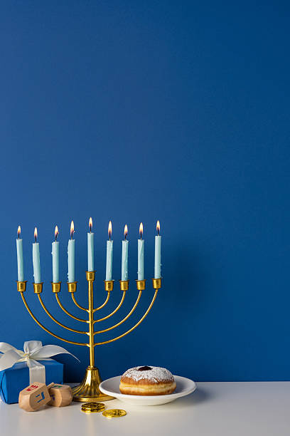 istock Colorful vertical Hanukkah scene featuring a gold menorah with lit candles, a doughnut on a plate, a dreidel, and wrapped gifts against a deep blue wall 2247028504