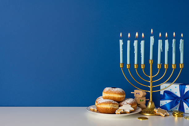 istock Festive Hanukkah scene features a glowing menorah, lit candles, powdered donuts, a wrapped gift, and dreidel tokens on a blue backdrop, conveying warmth, tradition, and celebration 2247028505