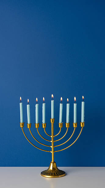 istock A vertical gold menorah stands on a white surface against a deep blue background, capturing a festive Hanukkah mood with lit candles and a warm, sacred glow 2247028510