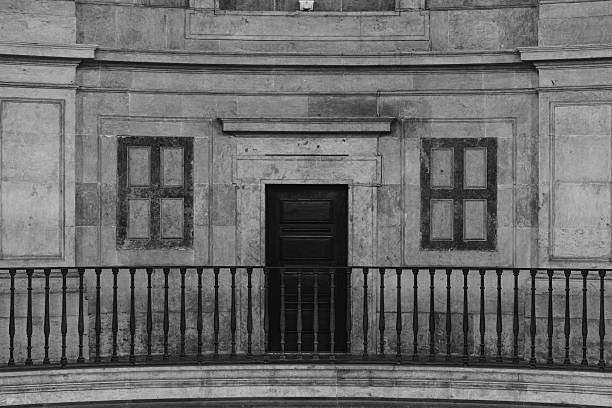Architectural detail of a symmetrical stone facade with a dark wooden door and balcony railing, captured in black and white