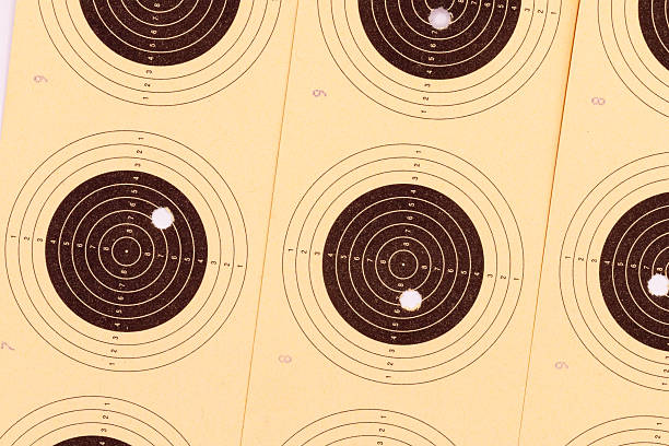 A top-down, close-up shot of multiple shooting targets. The targets have concentric black rings with numbered scoring zones on a light yellow paper. Some targets show bullet holes.