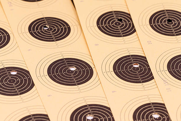 A close-up, angled view of several used paper shooting targets, arranged in overlapping layers. The targets are light brown with concentric black circles and scoring numbers.