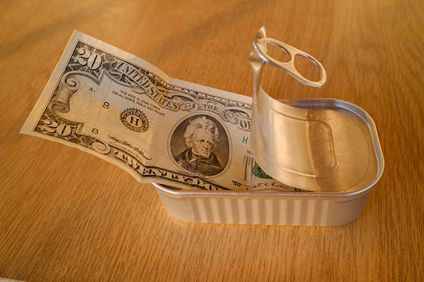 Twenty dollar bill trapped inside an open tin can on a wooden table, visualizing rising food prices, cost of living, savings under pressure, inflation and household expense strain