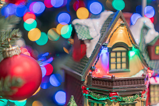 A charming gingerbread house, adorned with a red door and white window frames, is displayed in the foreground amidst twinkling Christmas lights in hues of blue, green, and red A large vibrant bauble
