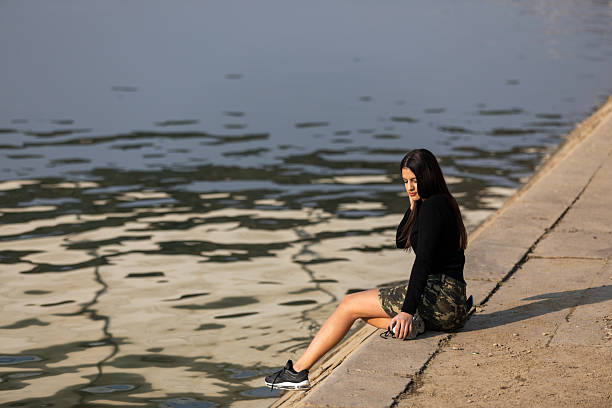 Young woman sitting at a riverbank, looking down in quiet reflection during a sunny afternoon, capturing solitude, contemplation and gentle urban summer calm