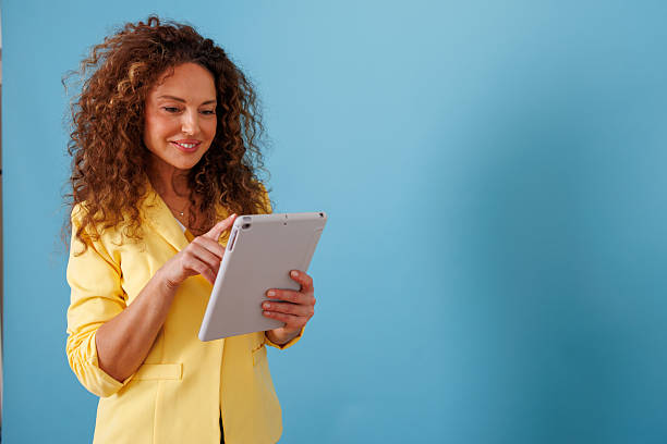 istock Businesswoman using digital tablet with yellow blazer on blue background while smiling and working 2247049341