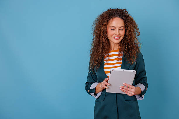 istock Cheerful young woman with curly hair showcasing her creative digital business solution on a tablet with a blue backdrop 2247050121