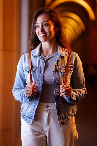 A cheerful young woman wears a denim jacket, gray shirt, and light pants, smiling while holding backpack straps in a warmly lit hallway; Ideal for lifestyle, fashion, travel, and confidence themes