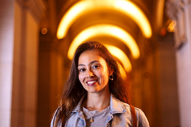 A cheerful young woman in a denim jacket poses in a warmly lit hallway with glowing arches, conveying confidence, friendliness, and contemporary style suitable for fashion, lifestyle, and portrait