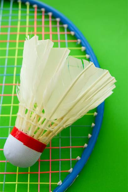 A high-angle flat lay photograph featuring a feather shuttlecock and the top of a badminton racket resting on a vibrant, uniform green background. The white shuttlecock and the blue racket frame create a dynamic visual contrast against the bold color, suggesting energy and movement. This image is excellent for themes related to badminton training, fitness, active lifestyles, and sports competition, emphasizing the core equipment needed for the game.
