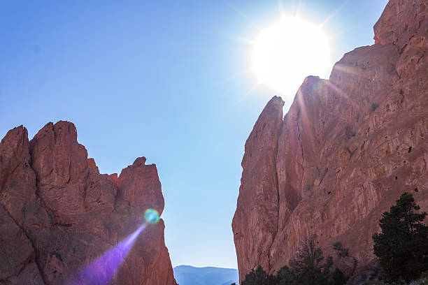 Sunlight streams over towering red rock formations in Colorado, casting dramatic shadows and highlighting the rugged texture against a clear blue sky.