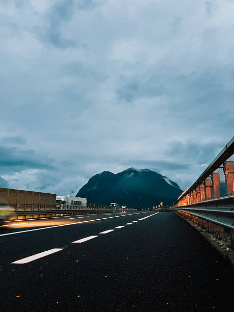 Empty highway disappearing into distance towards mountains under cloudy sky at dusk, with truck driving in distance and lights of passing car creating light trails. Asphalt road, mountain silhouettes