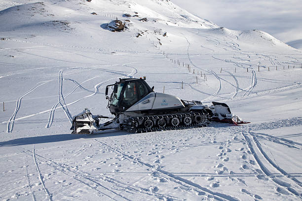Red snow groomer vehicle stands parked on snowy mountain ridge under dramatic cloudy sky. Snowcat captured against scenic alpine backdrop with snow-covered peaks