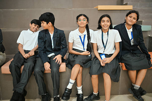 School students sitting close together on tiered steps inside a school wearing uniforms. The students appear relaxed and cheerful; two boys on the left share a playful moment, while the others smile warmly toward the camera.